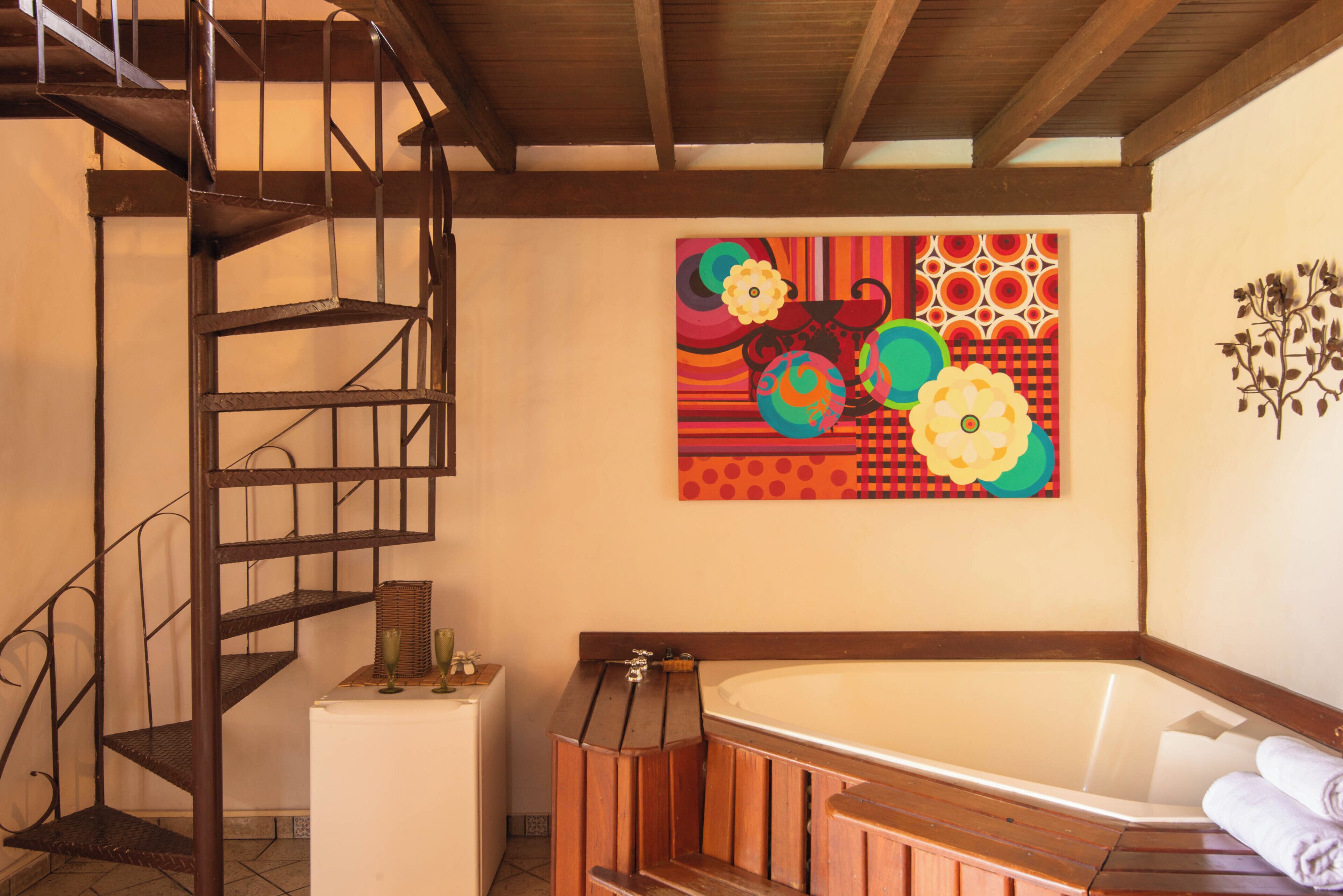 A bathroom with a wooden bathtub, a metal spiral staircase, and a colorful abstract painting on the wall.
