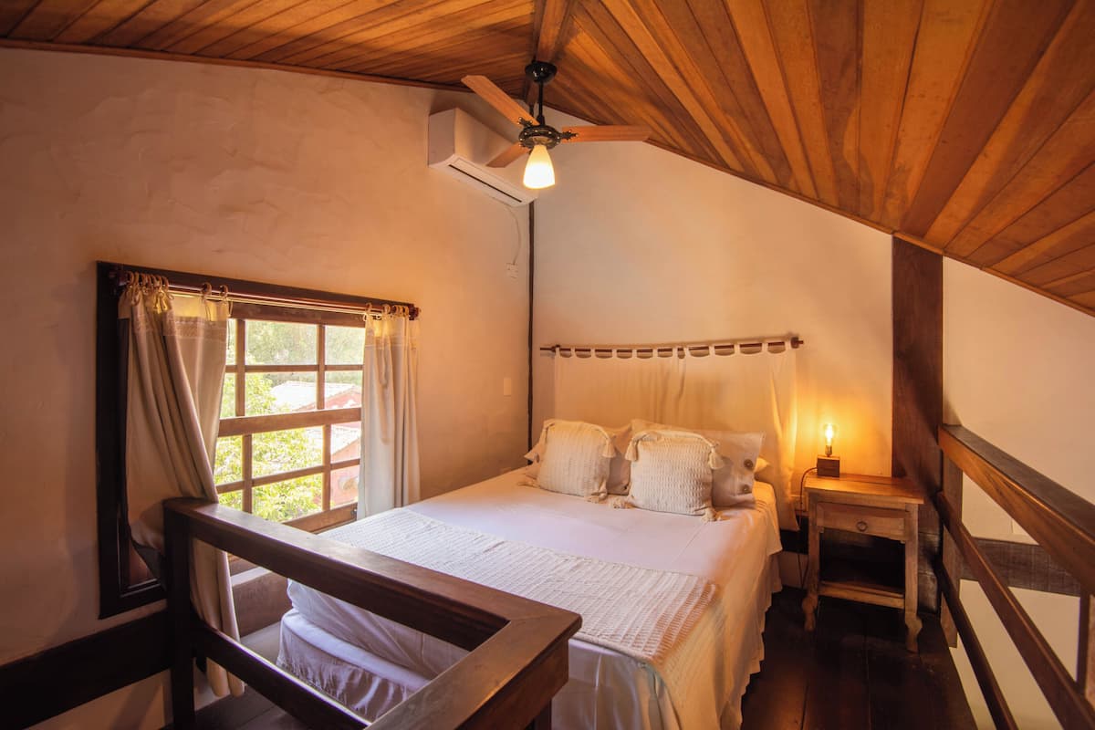 Ein Schlafzimmer mit Holzbalkendecke, einem Bett mit weißer Bettwäsche, einem Nachttisch mit Lampe und einem Fenster mit Vorhängen.