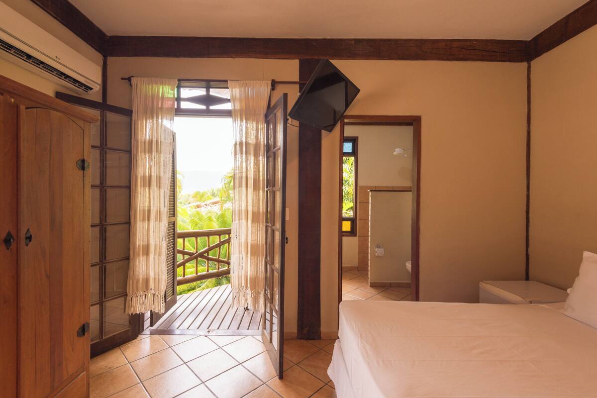 Ein Schlafzimmer mit Bett, einem Holzschrank, einem Badezimmer mit Toilette und einem Balkon mit Blick auf Grünflächen.