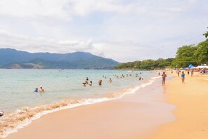 A beach with people swimming and walking along the shore, mountains in the background, and a clear sky. - Pousada Villaggio Assis (Ilhabela)