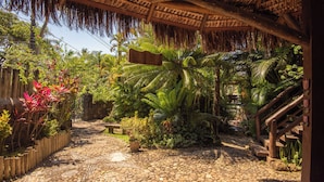 Un jardín tropical con un sendero de piedra, cerca de madera y una estructura con techo de paja.