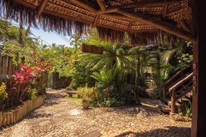 A tropical garden with a stone pathway, wooden fence, and a thatched-roof structure. - Pousada Villaggio Assis (Ilhabela)