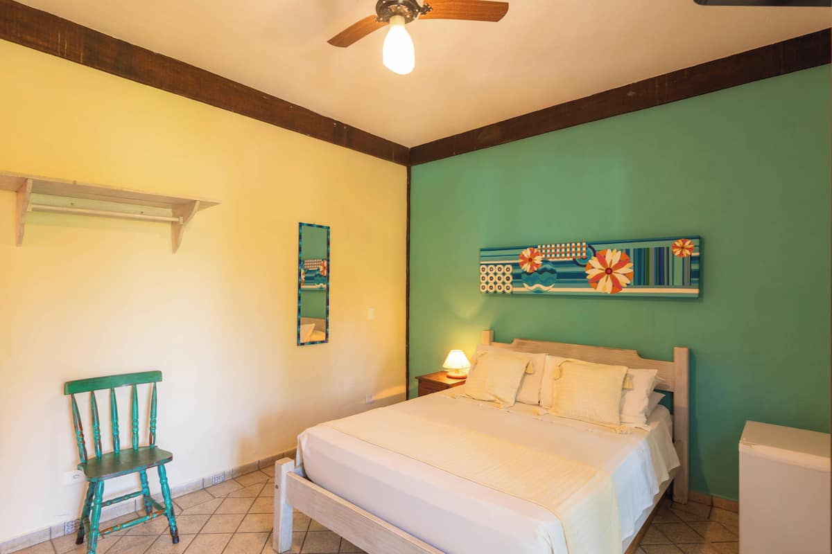 Ein Schlafzimmer mit einem Bett, einem Sessel, einem Nachttisch mit Lampe, einem Deckenventilator und einer Wand mit einem dekorativen Gemälde.