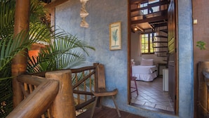 Una silla de madera en una habitación con piso de baldosas, un cuadro en la pared y una escalera que lleva a otra habitación.