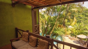 Un banco de madera con cojines, una barandilla de madera y vistas a un jardín tropical con palmeras.