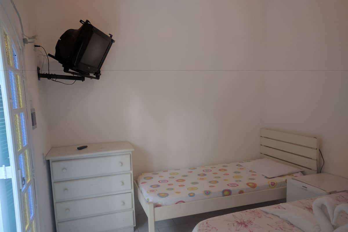 Um quarto pequeno e branco com uma cama de casal, uma mesinha de cabeceira, um armário e uma televisão fixada na parede.