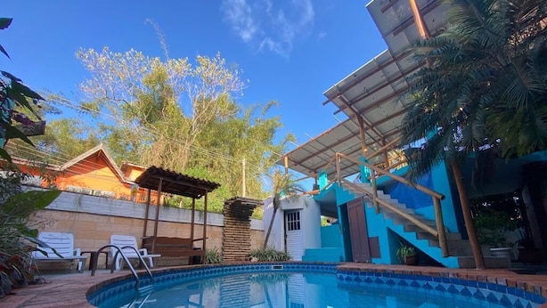 Una piscina con borde de ladrillo, una zona cubierta con escaleras y un edificio de fachada azul.