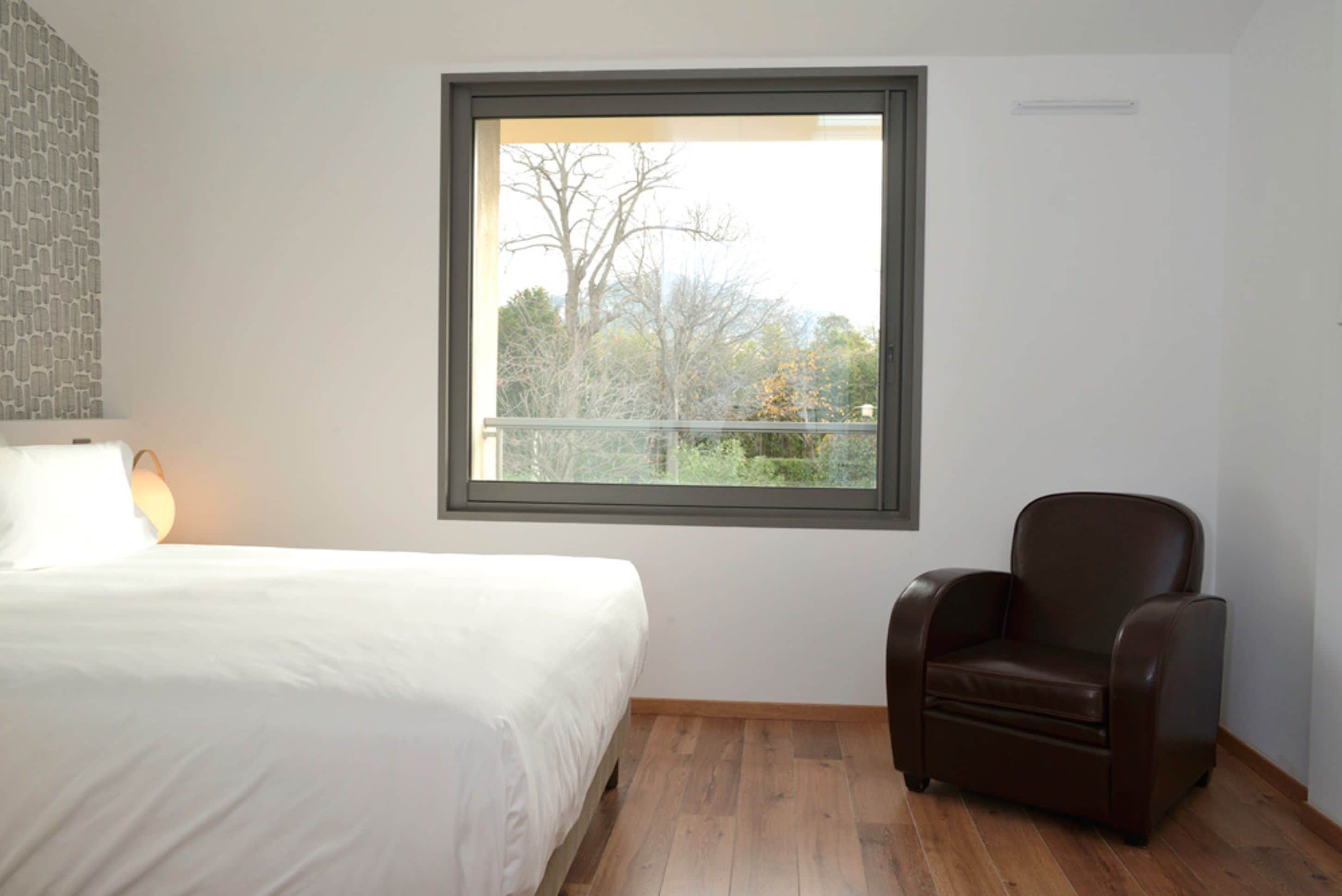 Un dormitorio con un ventanal grande, una cama, una silla y suelo de madera.