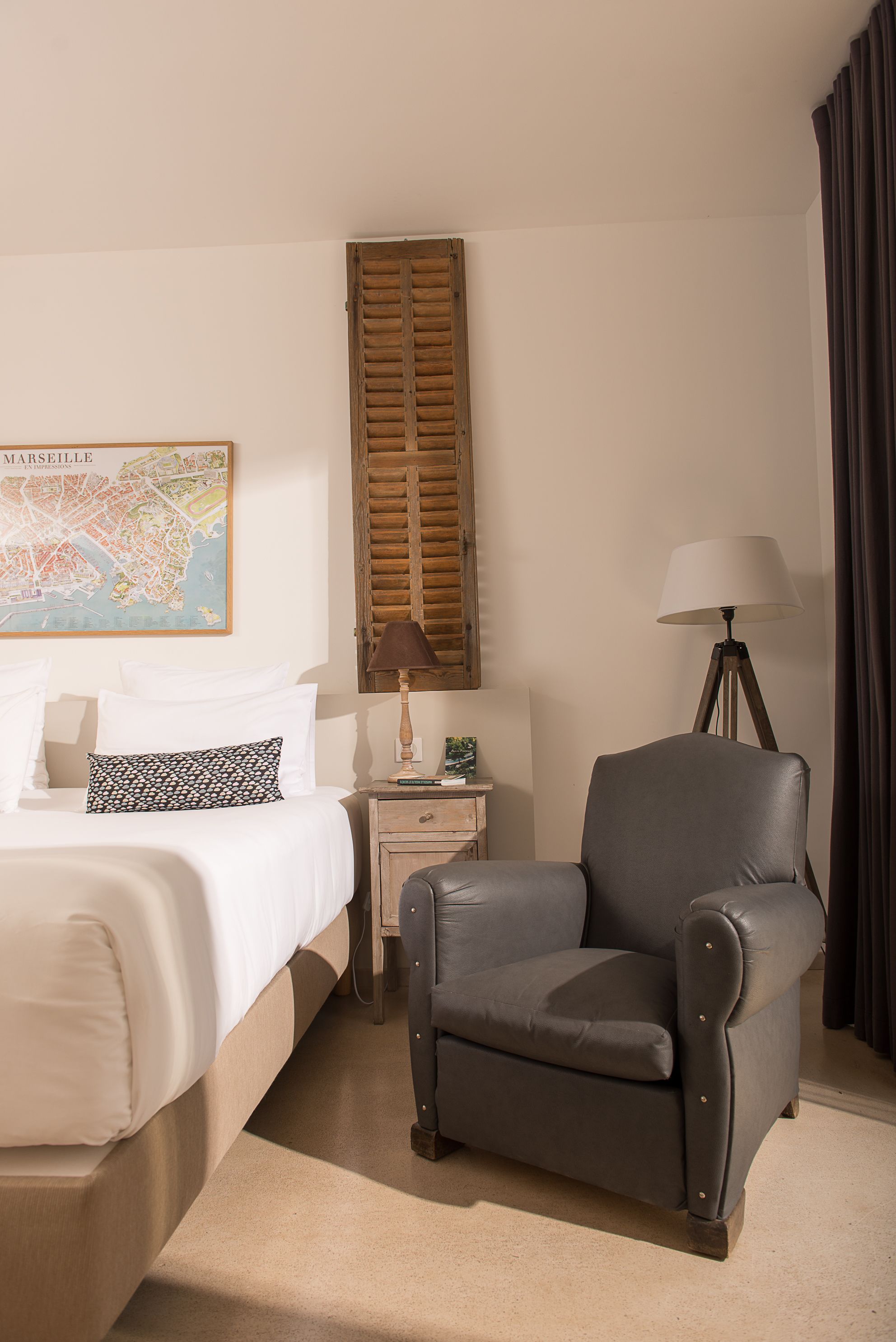 A hotel room with a bed, a grey armchair, a nightstand with a lamp, and a map on the wall.