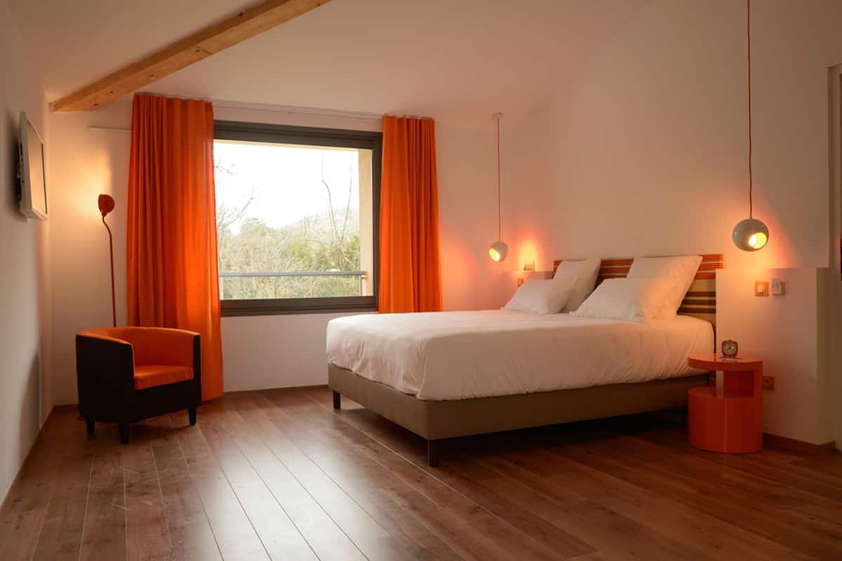 A bedroom with a bed, a chair, a window with curtains, and a bedside table with a lamp.