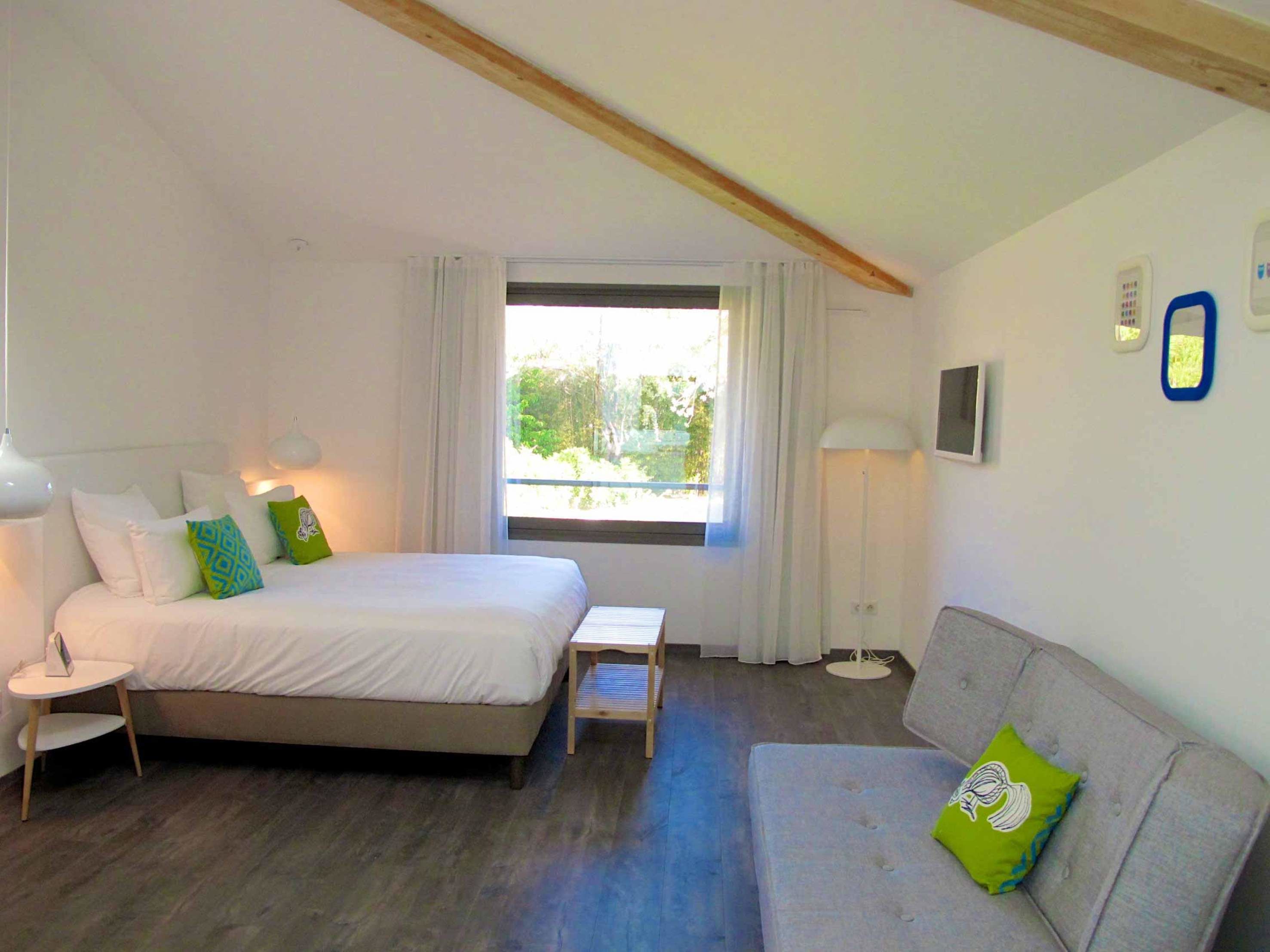 A bedroom with a bed, a bedside table, a lamp, a sofa, and a window with curtains.