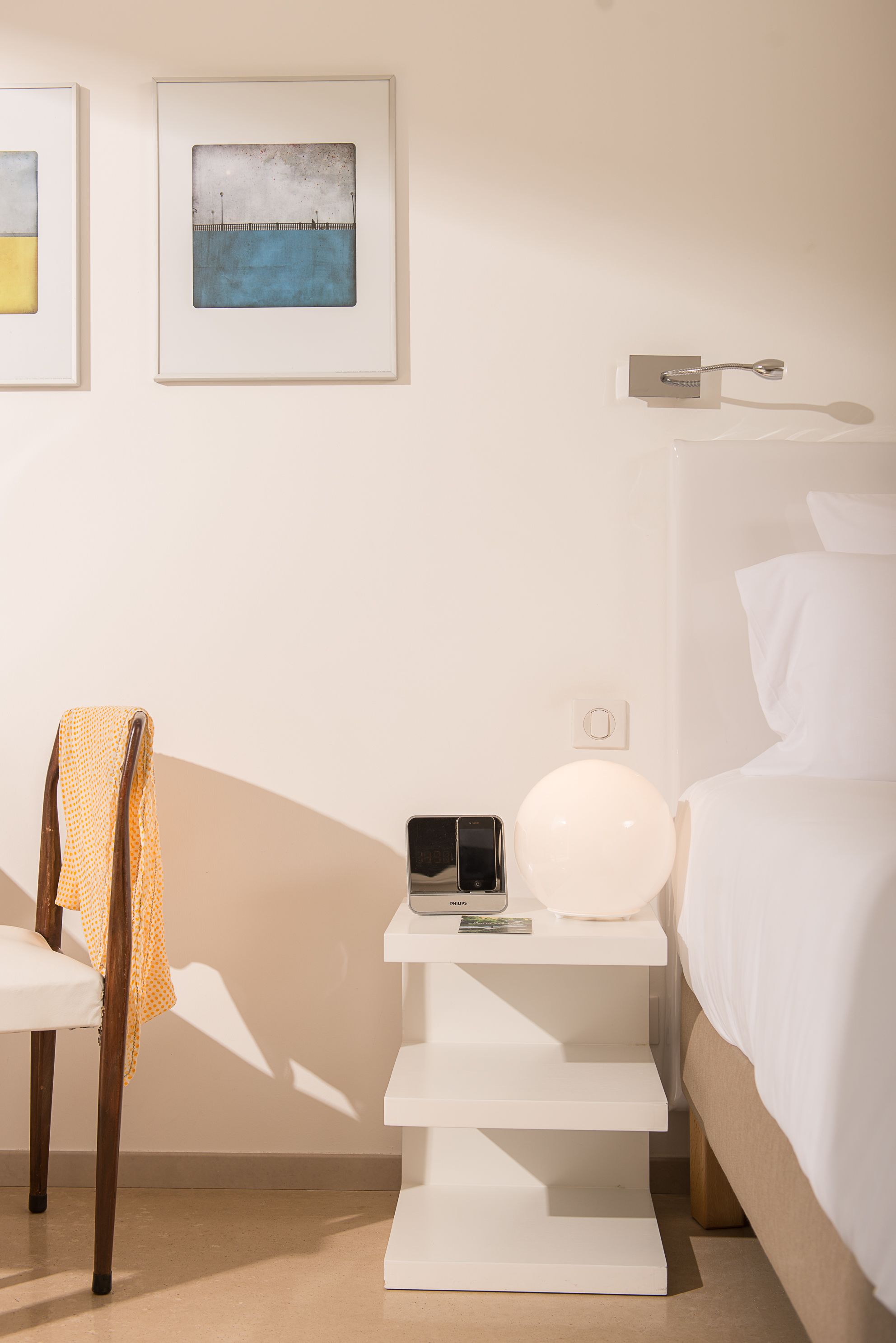 Habitación de hotel con cama, mesita de noche con lámpara, una silla y dos cuadros en la pared.
