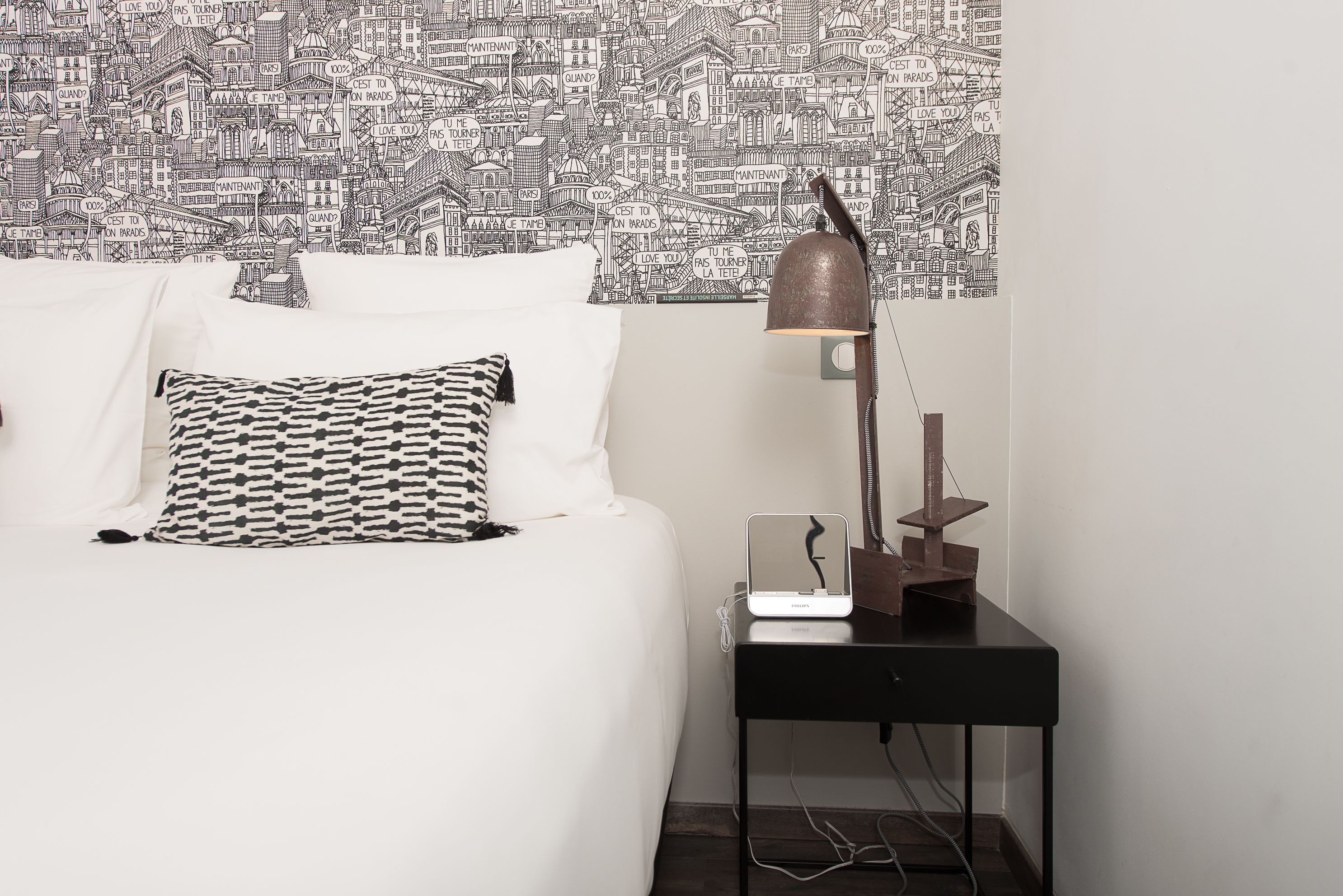 Una cama bien hecha con un almohadón estampado, una lámpara de noche y una mesita con una lámpara.