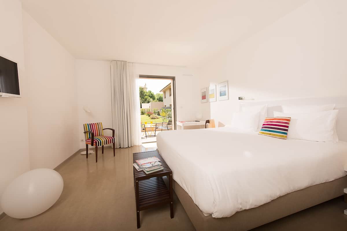 A hotel room with a large bed, a small table with magazines, a striped chair, and a view of the outside.