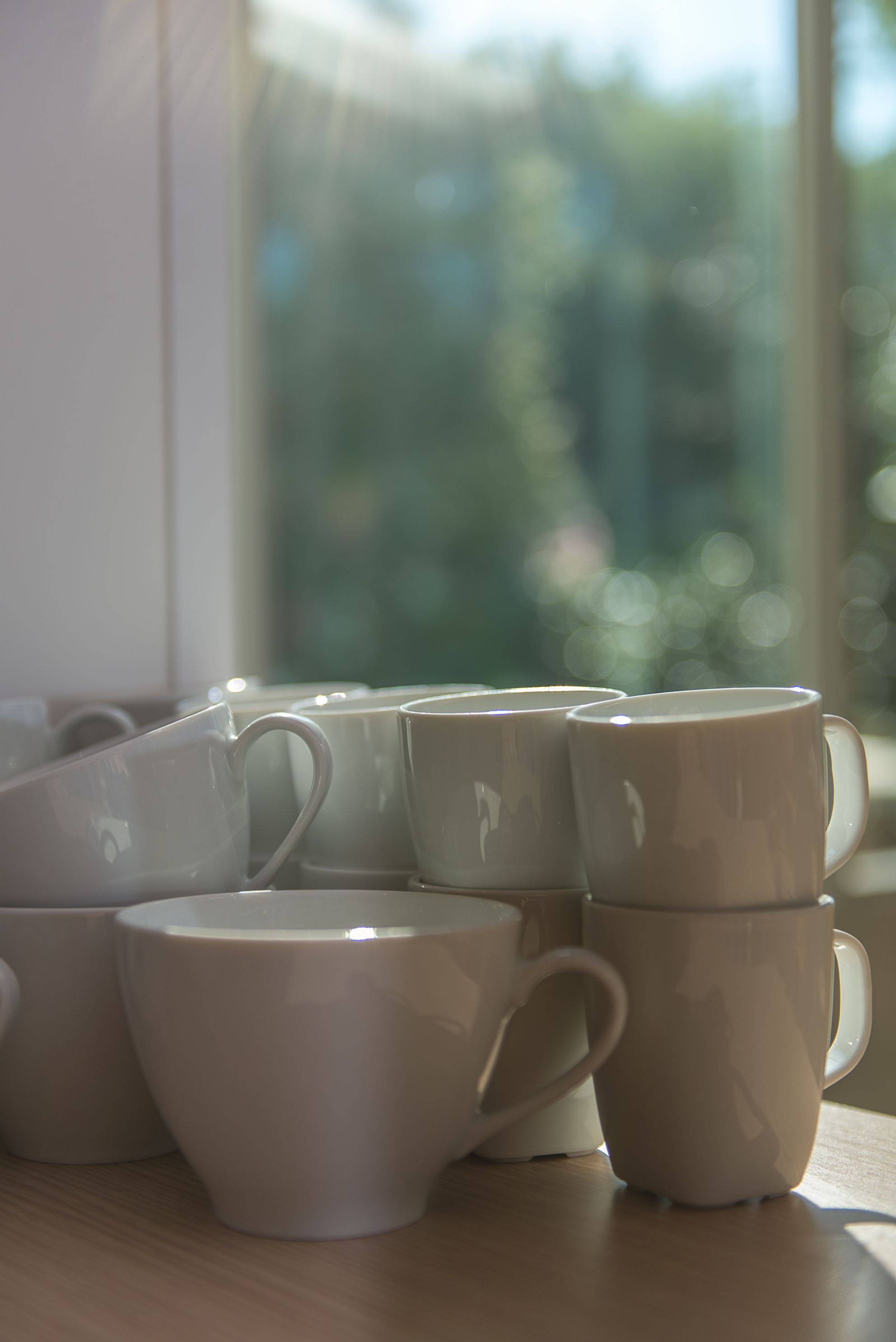 A stack of white ceramic mugs on a wooden table near a window.