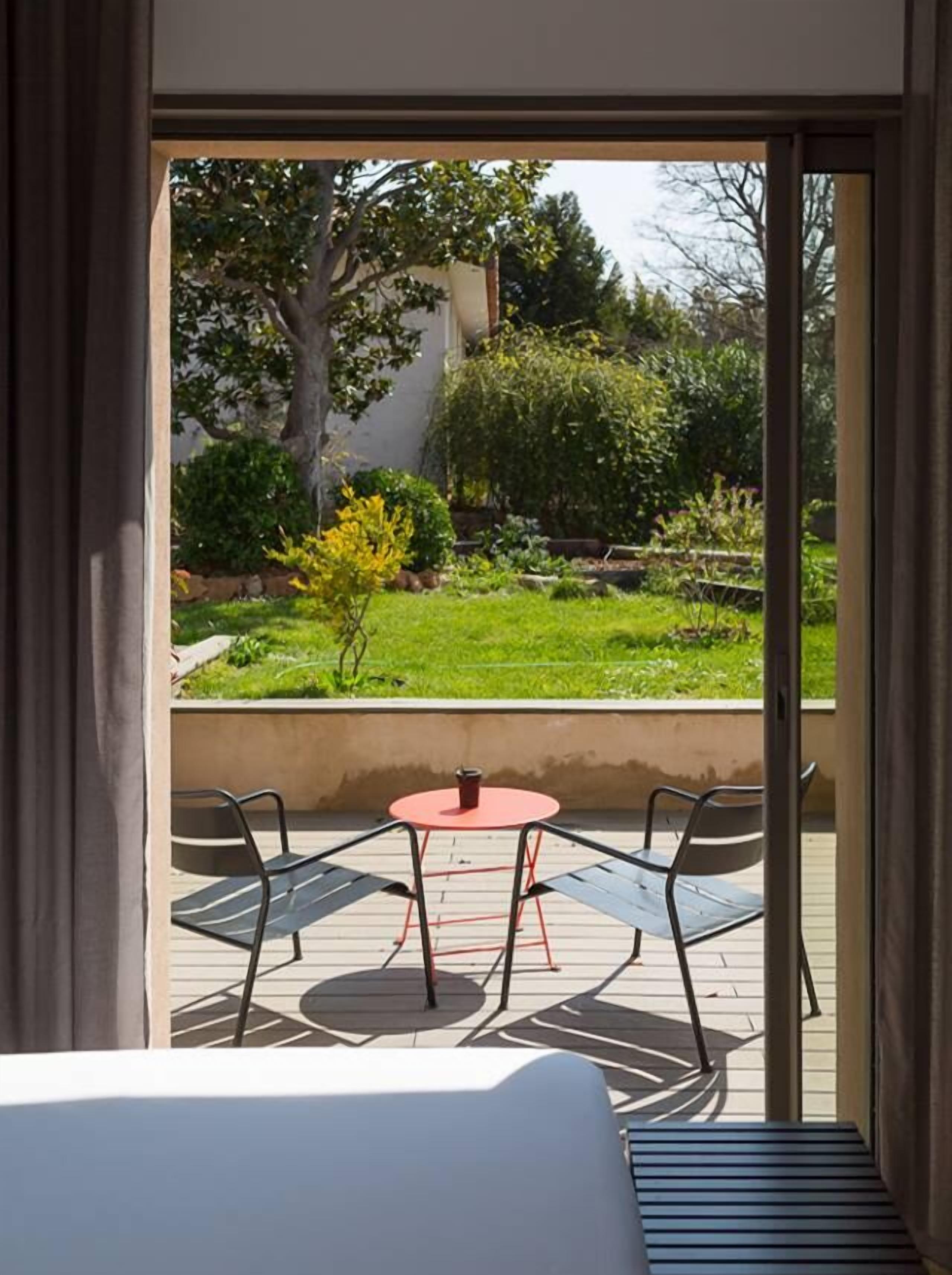 A room with a bed, a small red table, two chairs, and a view of a garden through a sliding door.