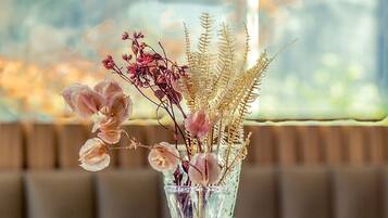 A table with a clear glass vase containing dried flowers and greenery, set on a table with a view of a cityscape through a window.