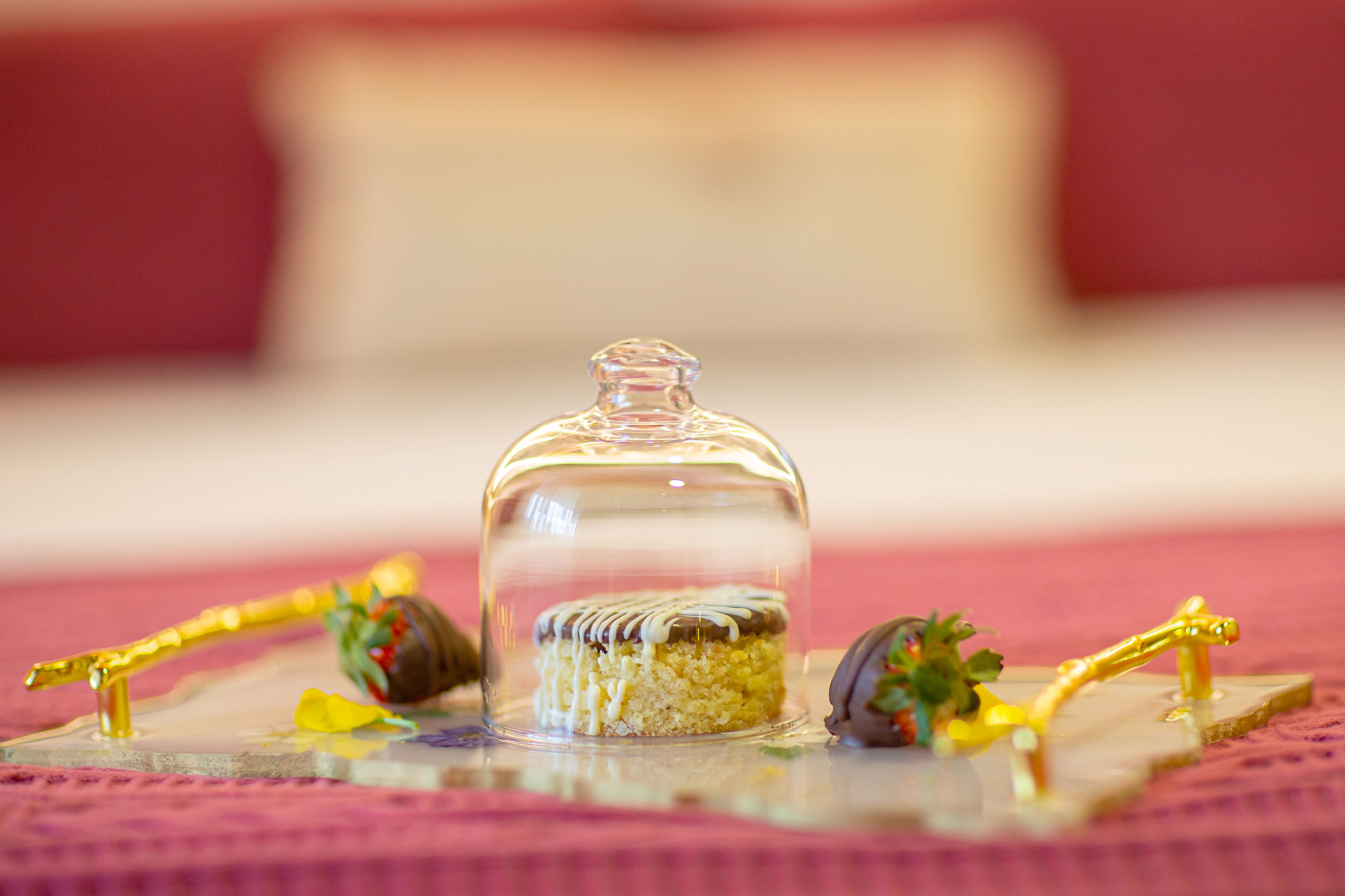Una mesa de postres con una campana de cristal, un pastel y fresas bañadas en chocolate sobre un mantel rosa.