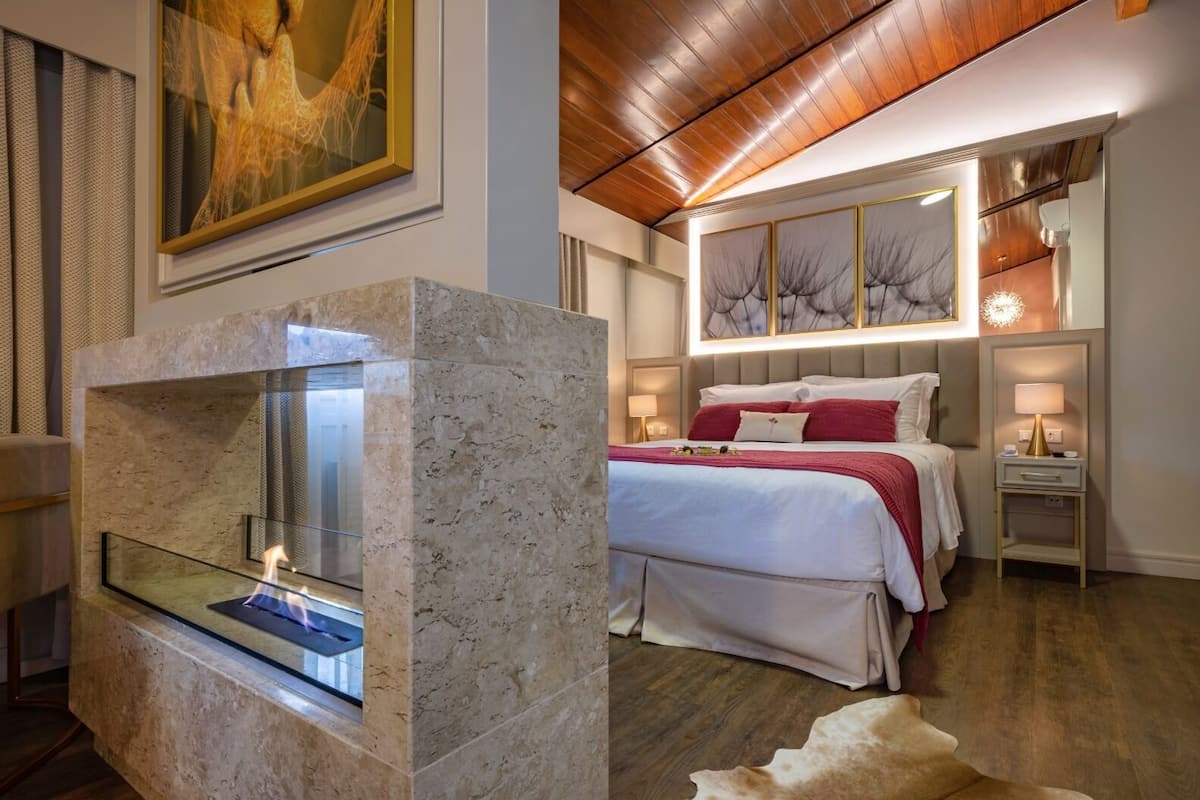 Ein modernes Hotelzimmer mit einem Marmor-Kamin, einem großen Bett mit roter Bettwäsche und einer Decke mit Holzbalken.
