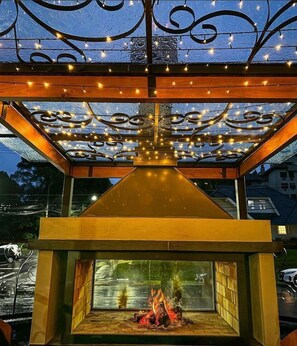 A modern outdoor fireplace with a glass enclosure, surrounded by a decorative metal structure with string lights. - Blumen Hotel Boutique (Canela)