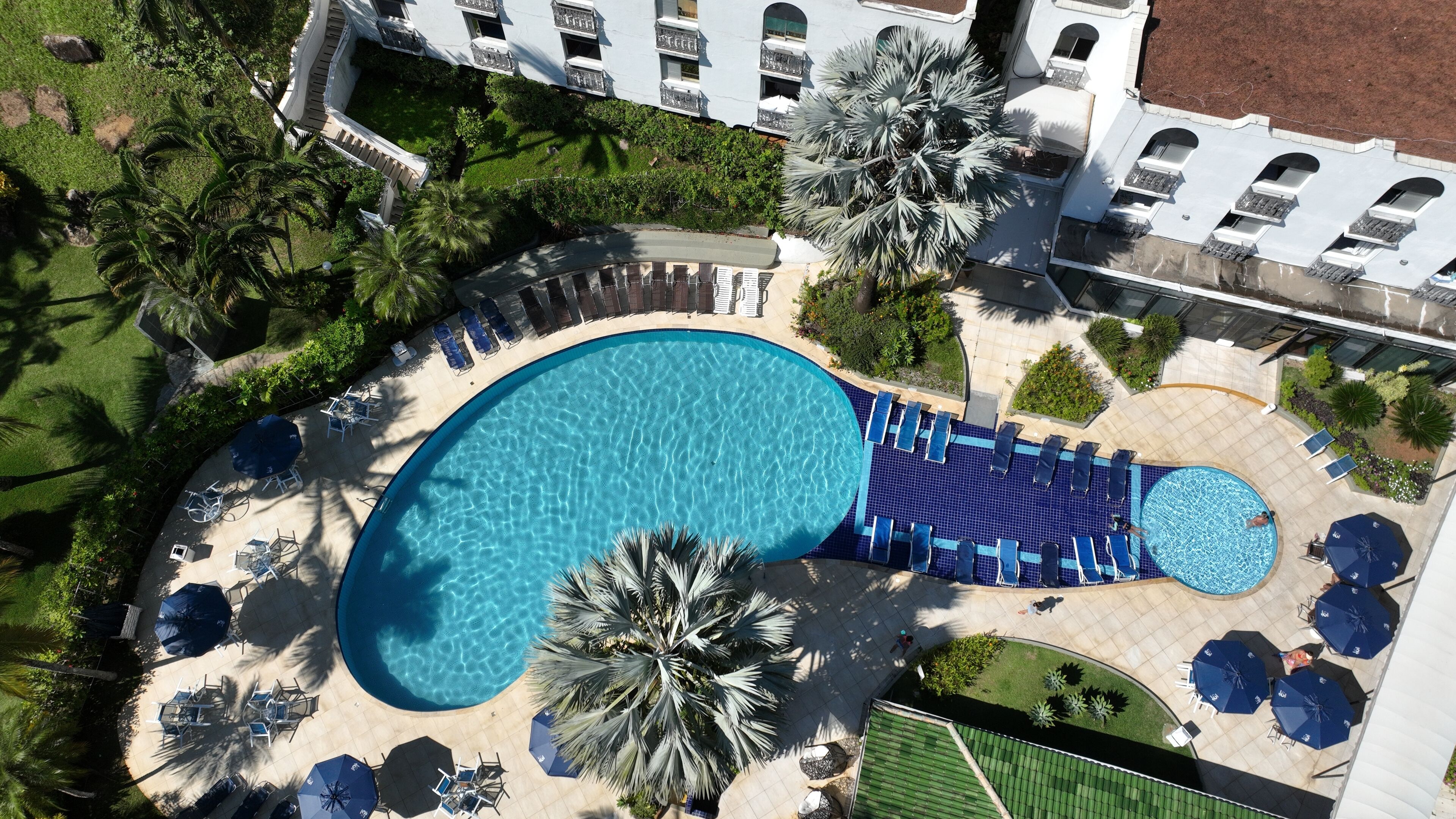 Vista aèria d'un complex turístic amb una gran piscina, gandules i palmeres.