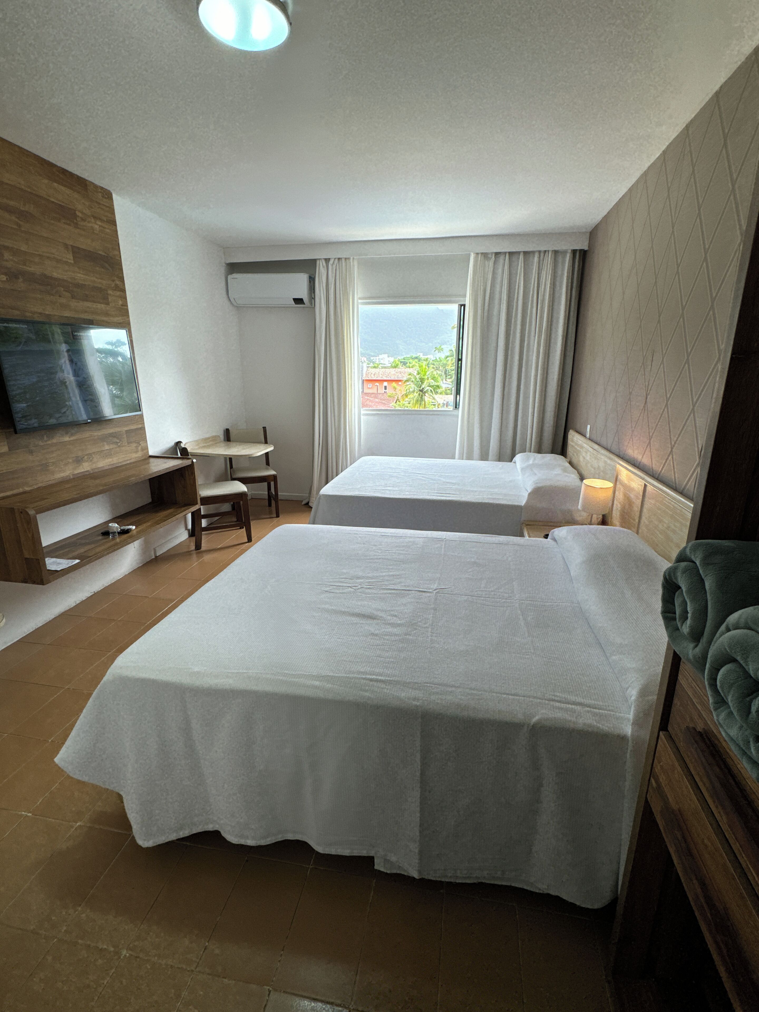 A hotel room with two beds, a TV mounted on the wall, a desk, and a window with curtains.