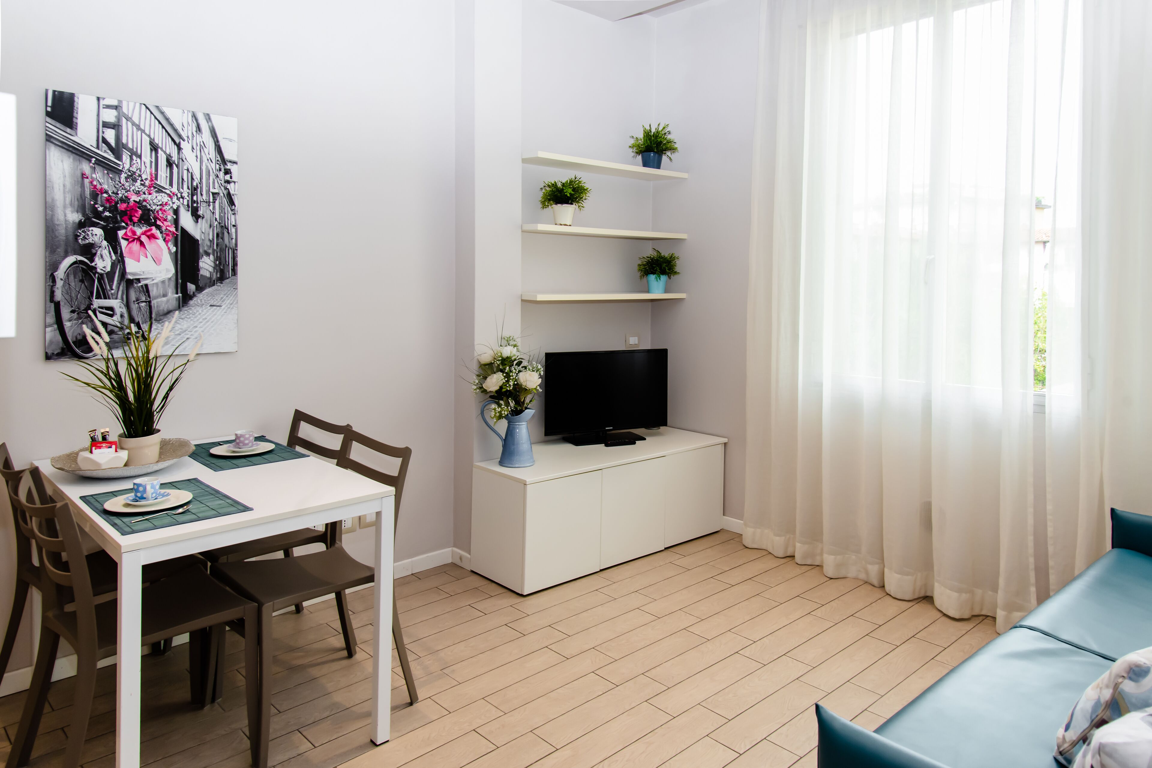 Una sala de estar moderna con comedor, un televisor de pantalla plana sobre un mueble blanco y una ventana con cortinas de gasa.