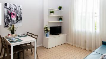 Una sala de estar moderna con comedor, un televisor de pantalla plana sobre un mueble blanco y una ventana con cortinas de gasa.