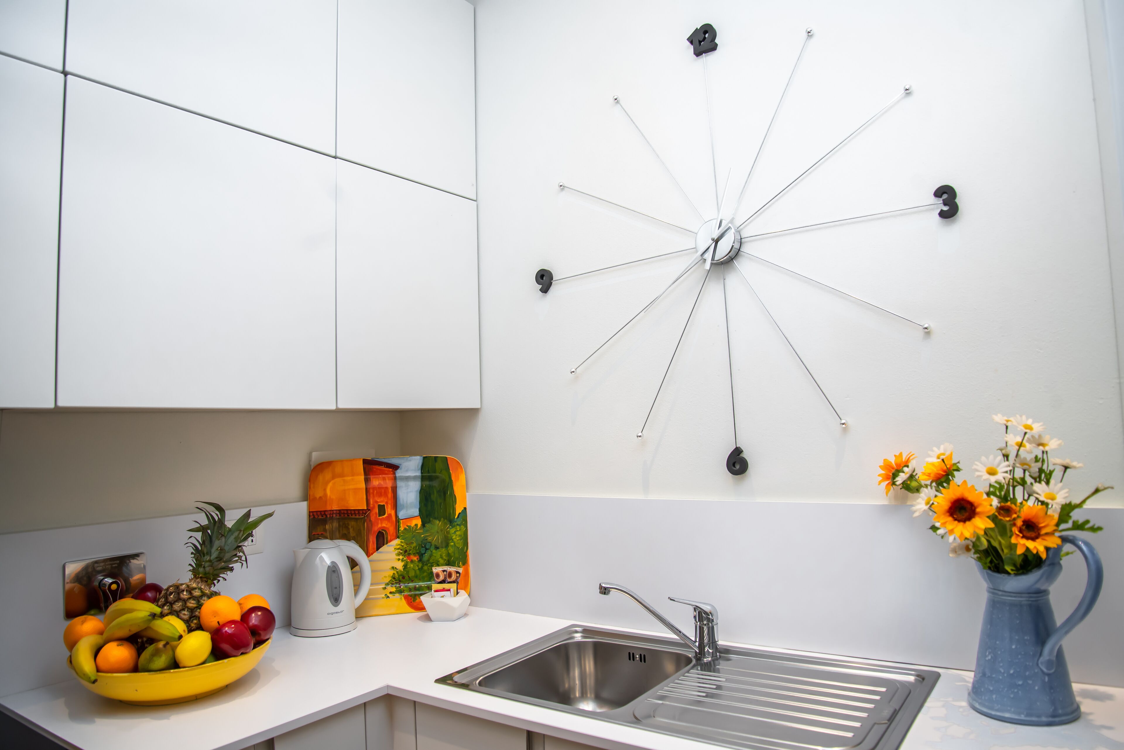 A kitchen with a fruit bowl, a kettle, a sink, and a wall clock.