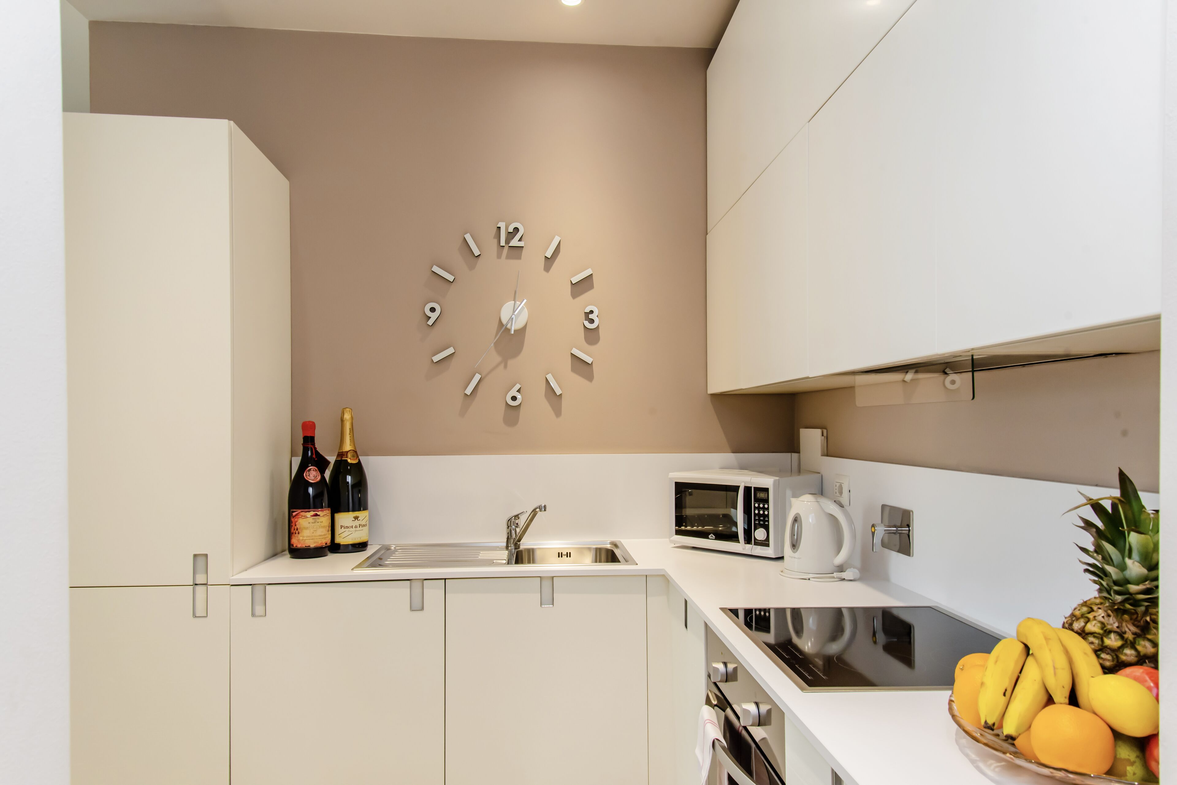 A modern kitchen with a wall clock, a sink, a microwave, and a fruit bowl.