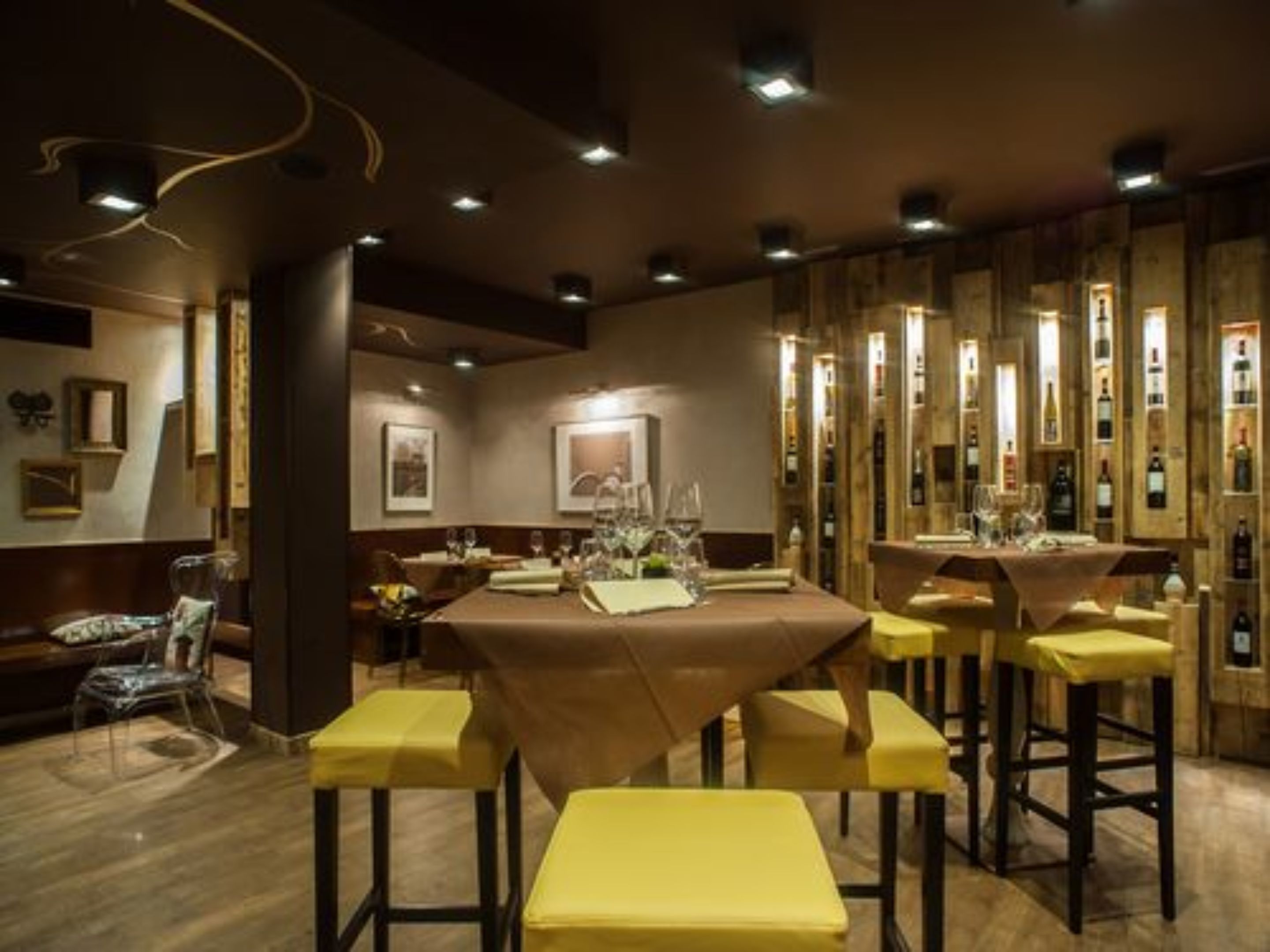 Un restaurante con una mesa larga, sillas amarillas y una estantería con botellas de vino al fondo.