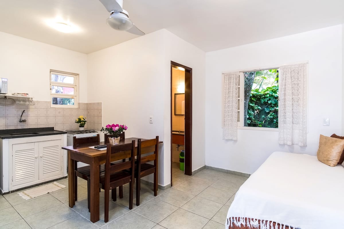 A compact room with a bed, a dining table with chairs, a kitchenette, and a window with curtains.