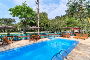 Una piscina con sillas de descanso y sombrillas, rodeada por una cerca azul y vegetación.