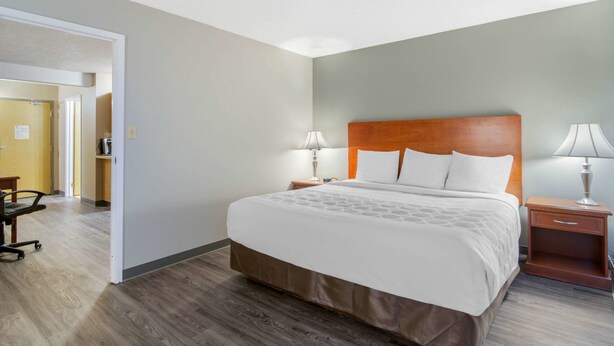 Quarto de hotel com uma cama grande, cabeceira de madeira, duas mesas de cabeceira com abajures, uma escrivaninha com cadeira e uma porta que dá para outro cômodo.
