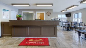 Um saguão de hotel com balcão de recepção, relógio, televisão e área de refeições com cadeiras e mesas.