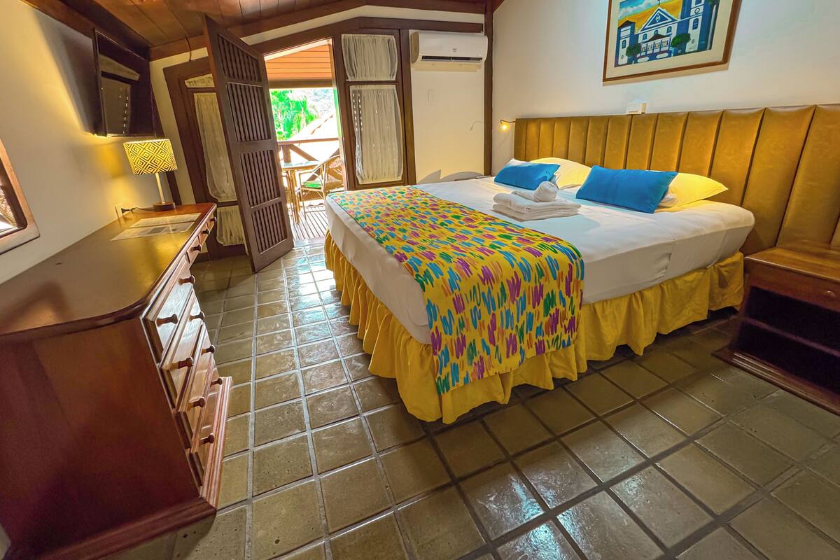 A hotel room with a bed, a wooden headboard, a nightstand, a desk, and a chair. There is a window with curtains, a door, and a painting on the wall.