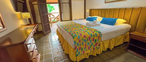 Habitación de hotel con cama, cabecera de madera, mesita de noche, escritorio y silla. Se ve una ventana con cortinas, una puerta y un cuadro en la pared.