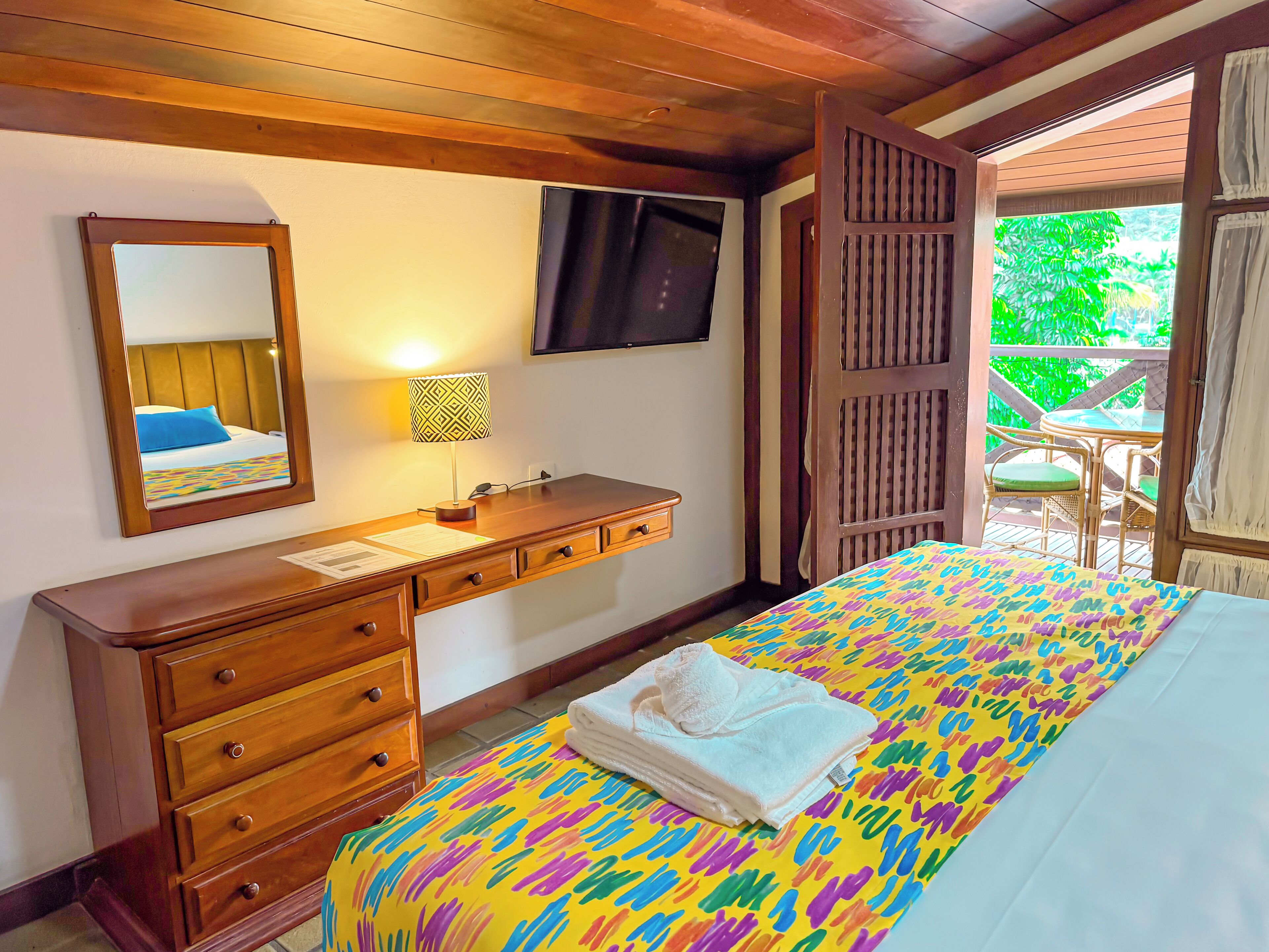 A hotel room with a bed, a wooden dresser, a mirror, a TV, and a balcony with a table and chairs.