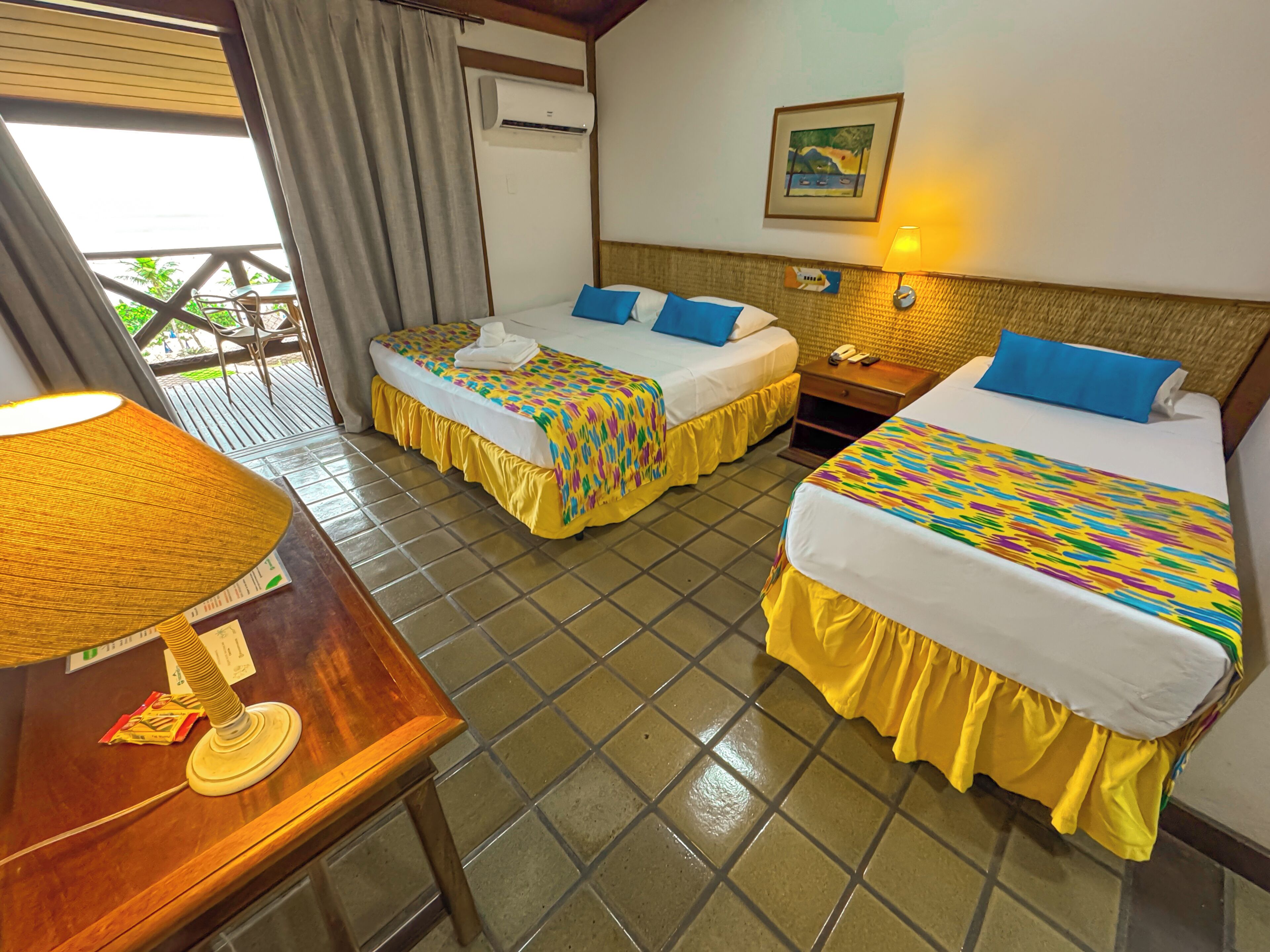 A hotel room with two beds, a wooden table, a lamp, an air conditioning unit, and a balcony with chairs.