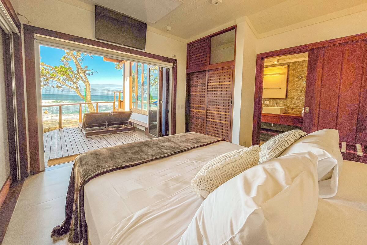 A bedroom with a large bed, a view of the beach, and a balcony with a chair.