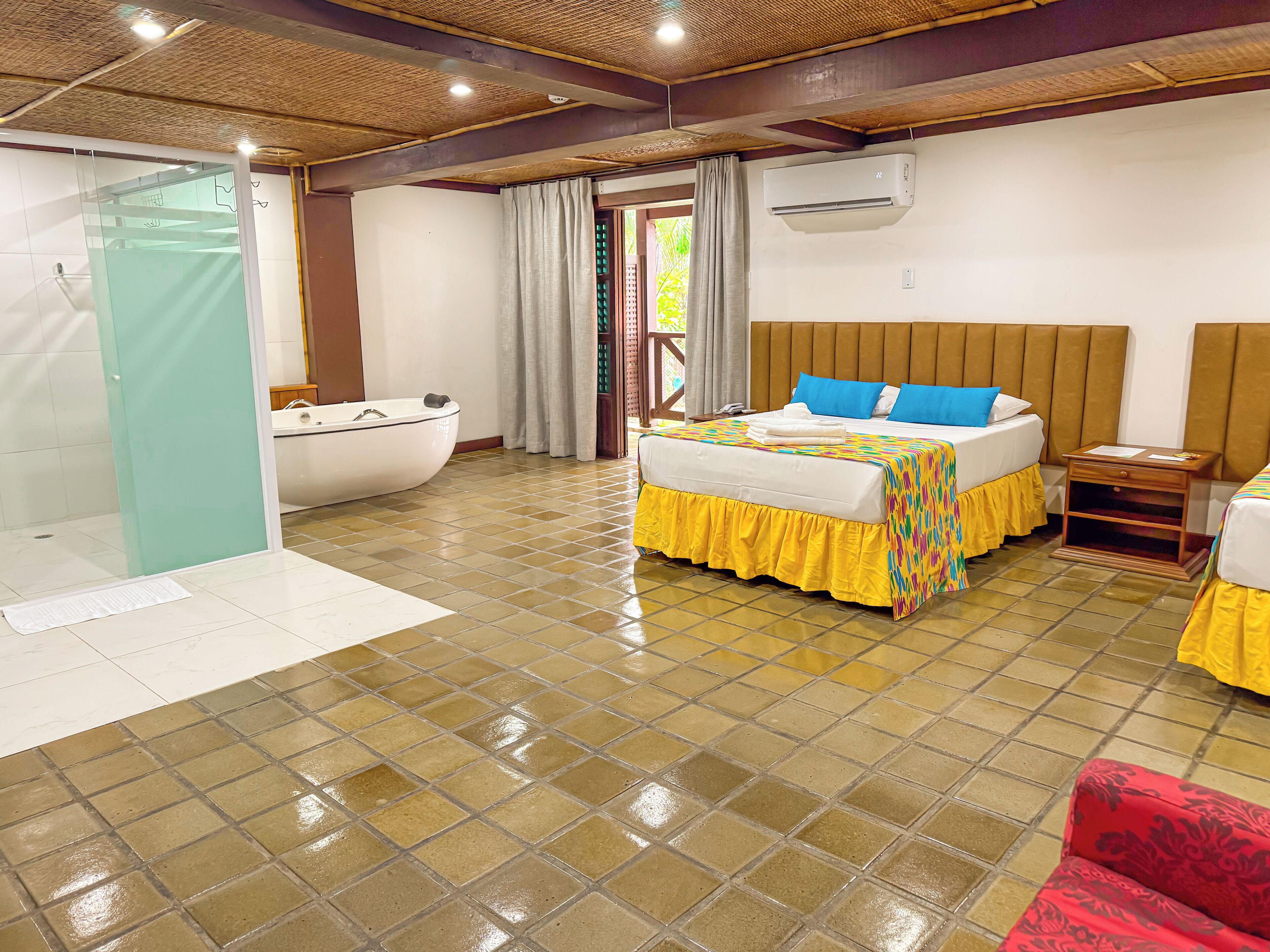 Una habitación de hotel con piso de baldosas, una cama con colcha colorida, cabecera de madera, mesita de noche y un baño con bañera.
