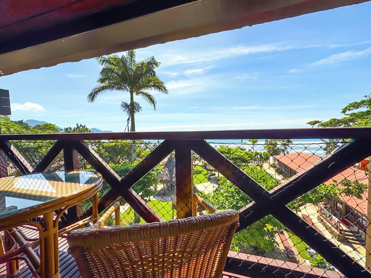 A balcony with wicker furniture, a glass table, and a view of a palm tree and buildings.