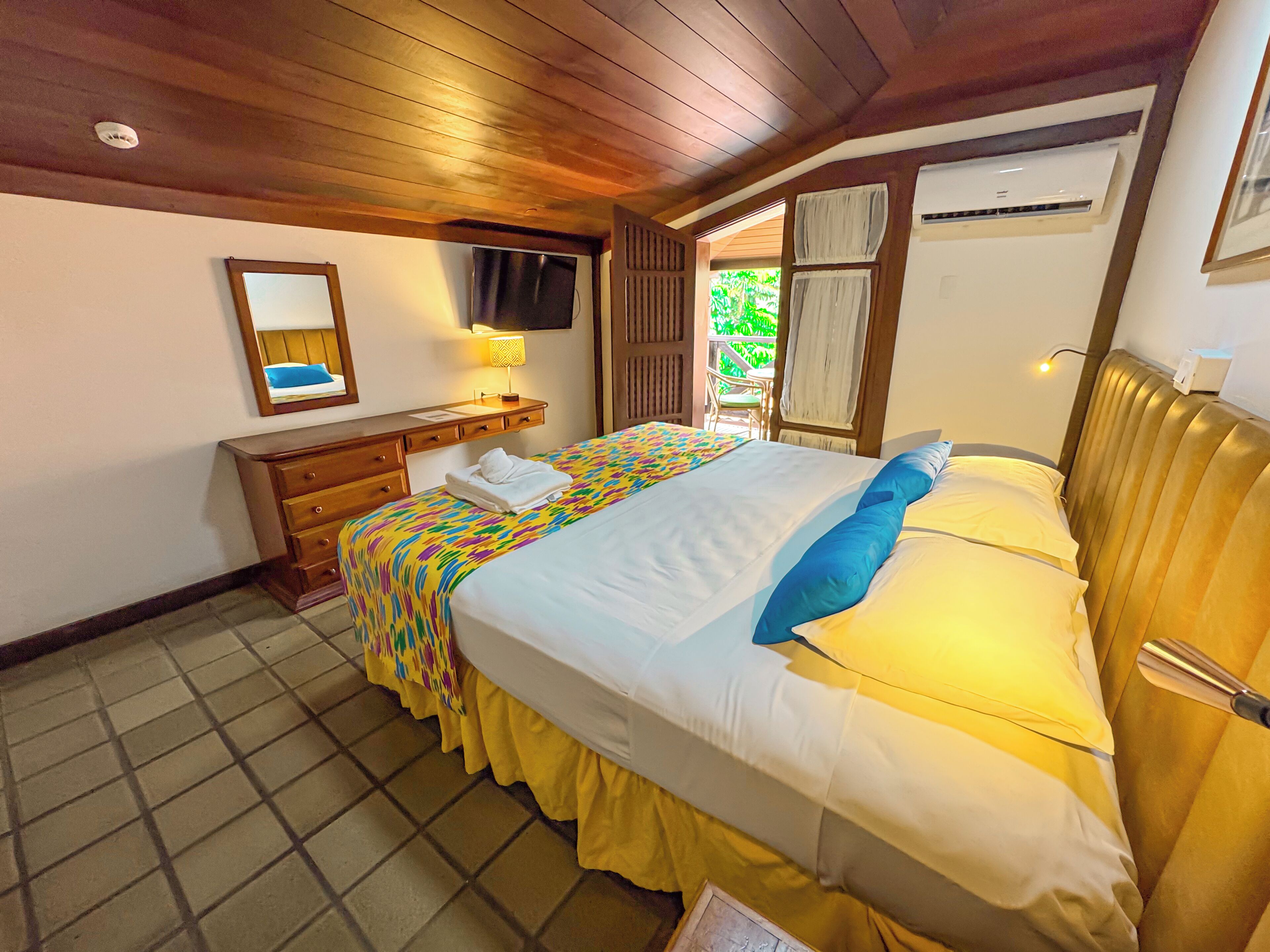 Quarto de hotel com cama, mesa de cabeceira, espelho, televisão e ar condicionado.