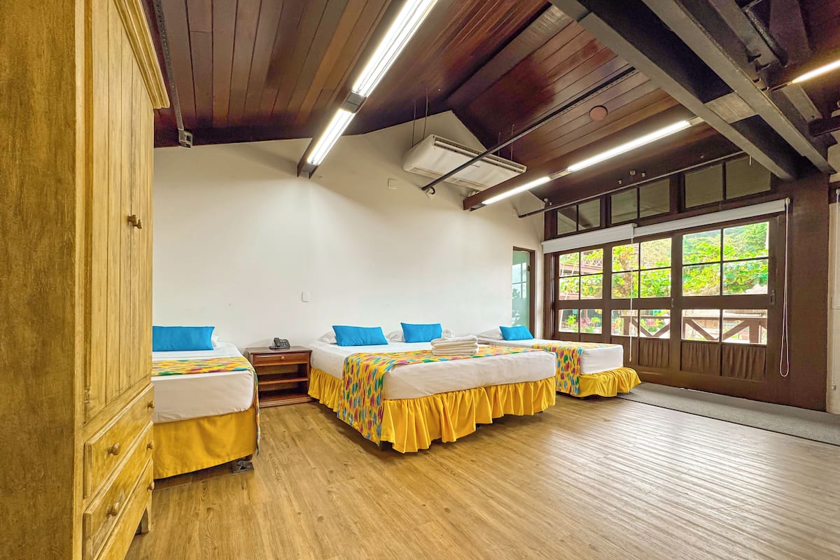 A hotel room with wooden floors, a large window with a view, and three beds with blue pillows.
