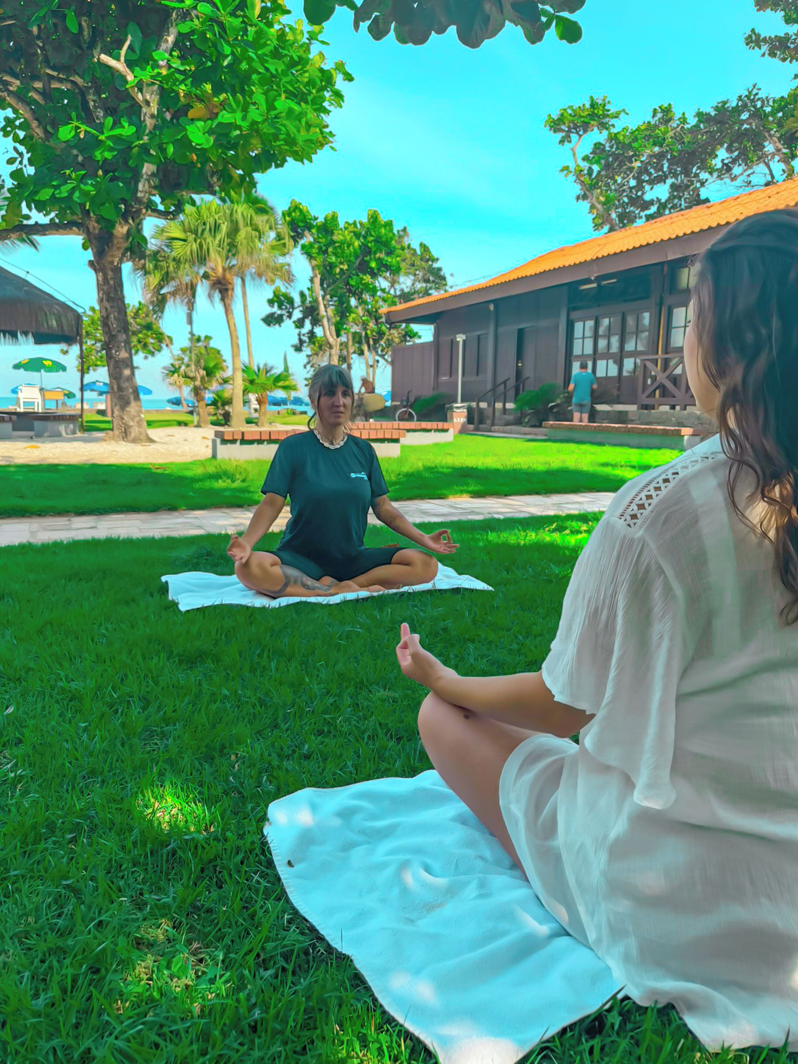 Due persone meditano in un prato con alberi e un edificio sullo sfondo.