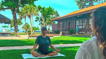 Due persone meditano in un prato con alberi e un edificio sullo sfondo.