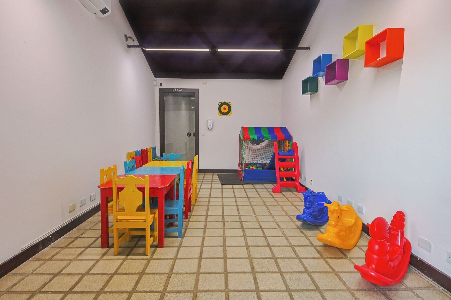 Espaço infantil com mesa e cadeiras, piscina de bolinhas e cubos de armazenamento coloridos na parede.
