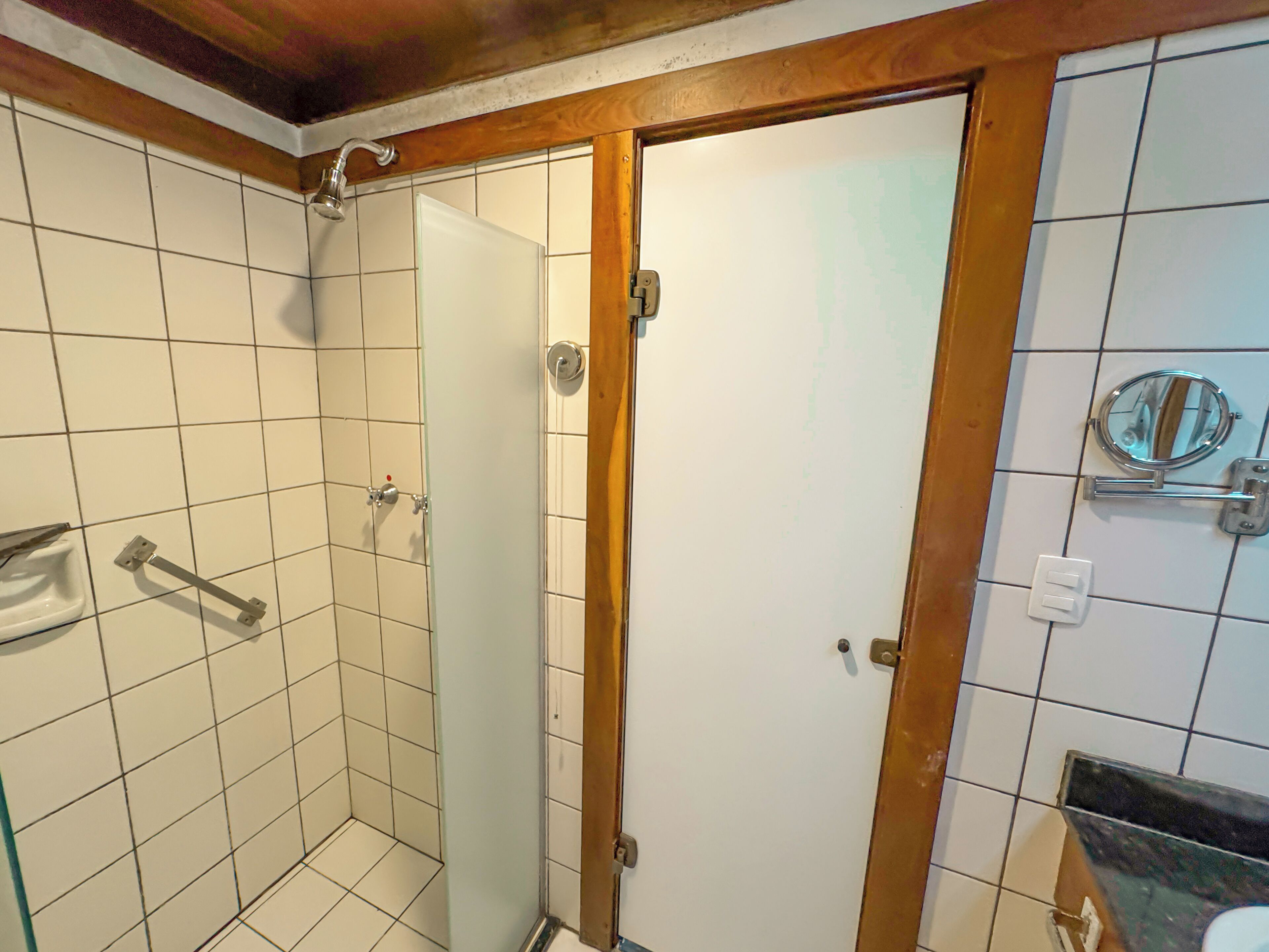A bathroom with white tiled walls, a glass shower enclosure, a wooden door, and a mirror with a magnifying glass.