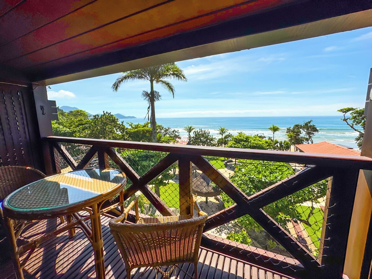 A balcony with wicker furniture and a glass table, overlooking a coastal landscape with palm trees and the ocean.