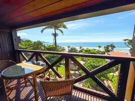 Un balcón con mobiliario de mimbre y una mesa de vidrio, con vista a un paisaje costero con palmeras y el océano.